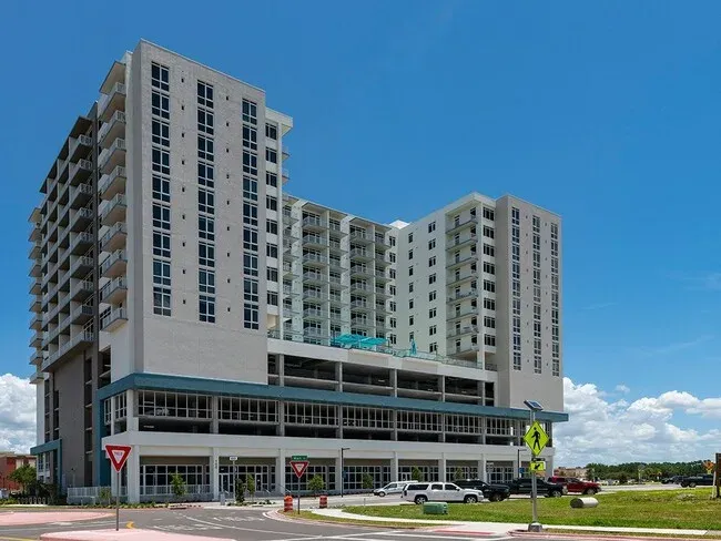 A contemporary multi-story apartment building featuring a mix of residential and commercial spaces, with a large parking area at the base and balconies on several floors. The structure is set against a clear blue sky, indicating a sunny day.