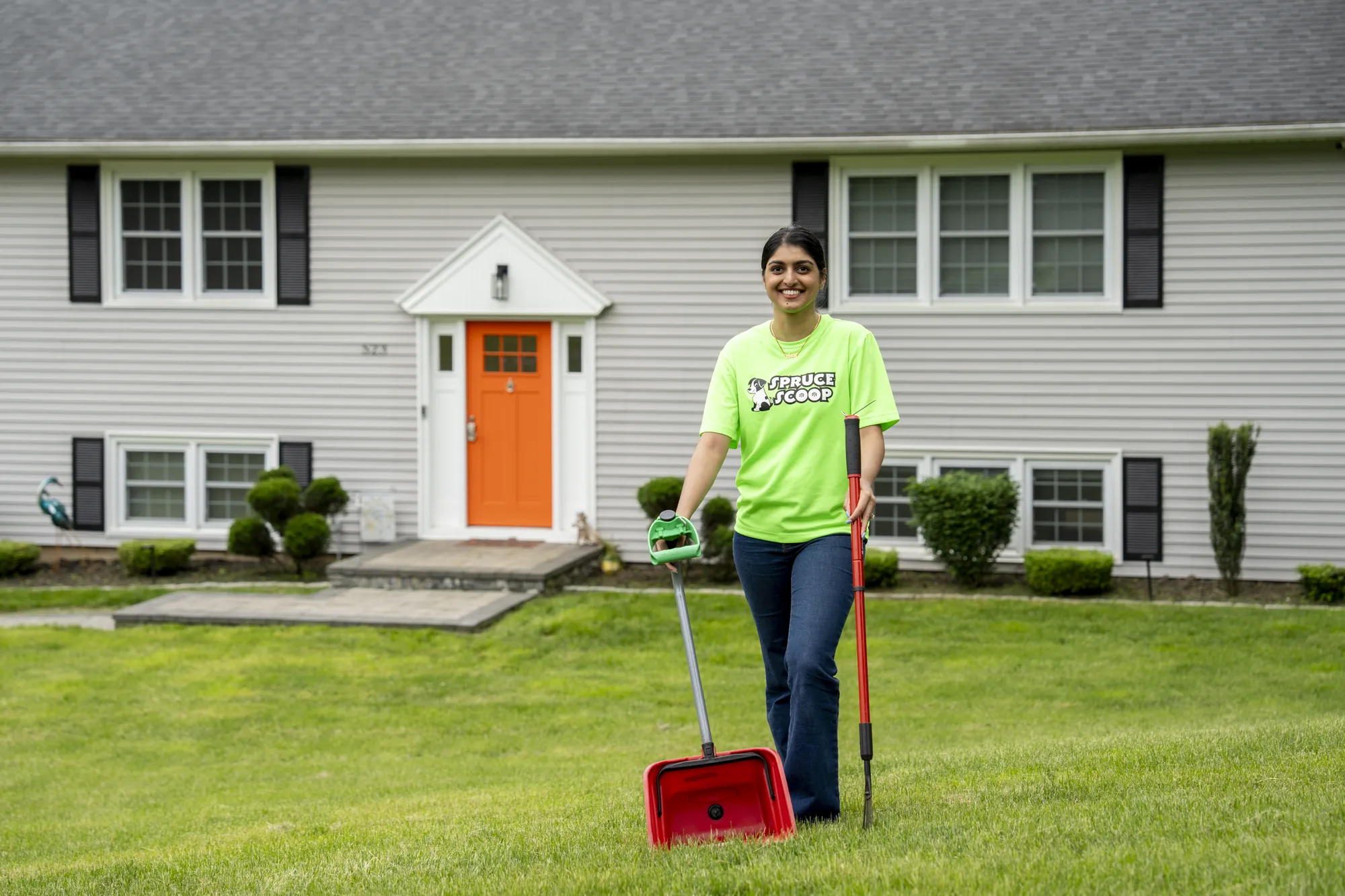 A Spruce Scoop team member with a rake and a red scooper pan on the front lawn of a house with an orange door